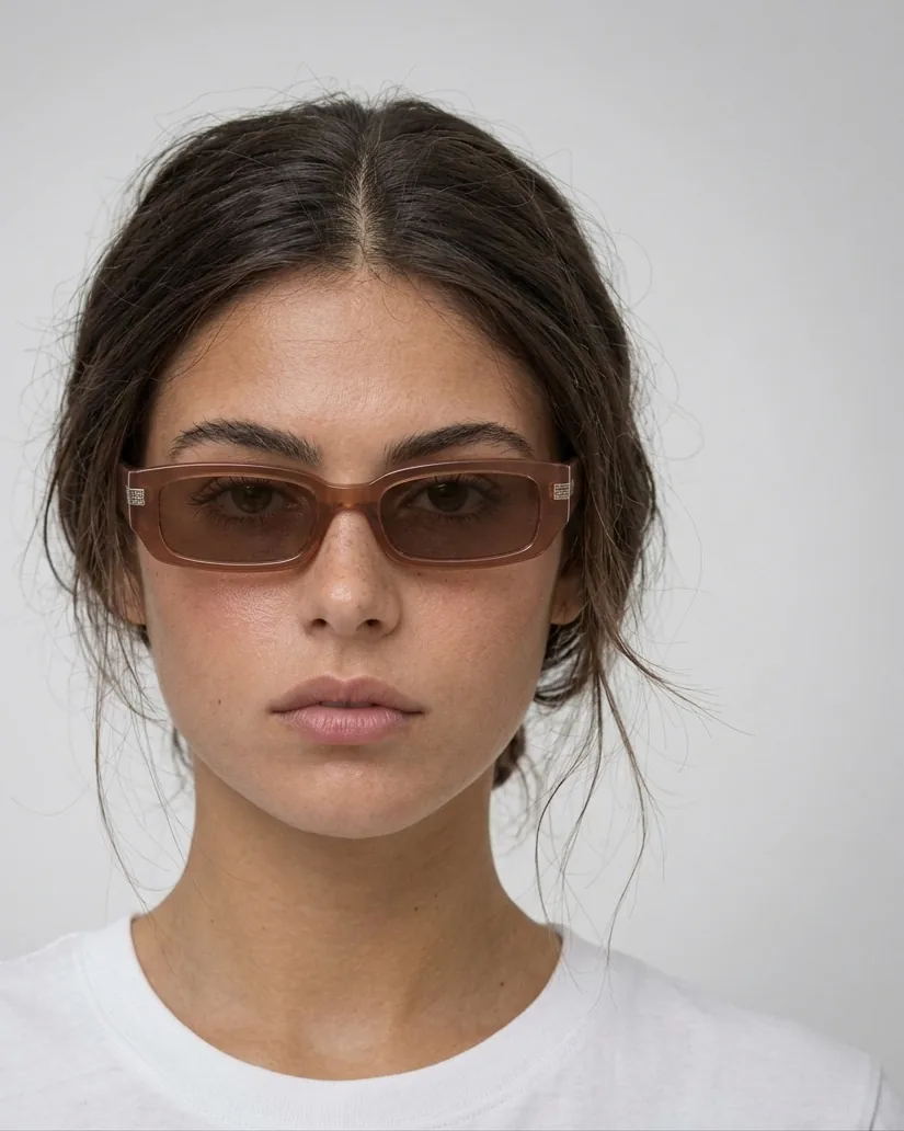 Primer plano de una modelo morena con gafas de sol de montura marrón translúcida y camiseta blanca, sobre fondo blanco