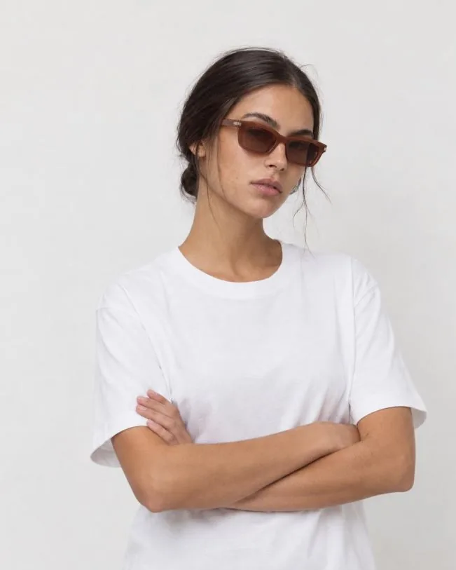 La misma modelo con gafas de sol marrones y camiseta blanca, de brazos cruzados, con la misma luz suave y fondo blanco